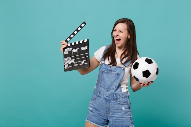 Laughing girl fanático del fútbol apoya al equipo favorito con una clásica película negra de balón de fútbol que hace una claqueta aislada en un fondo azul turquesa. Emociones de la gente, concepto de estilo de vida de ocio familiar deportivo.