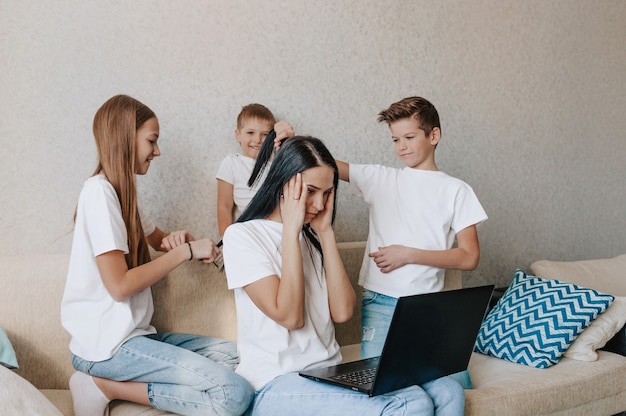 Laut verwöhnte Kinder verhindern, dass ihre Mutter ferngesteuert an einem Laptop arbeitet, während sie zu Hause auf der Couch sitzt. lenke deine Mutter ab, indem du an ihren Haaren ziehst