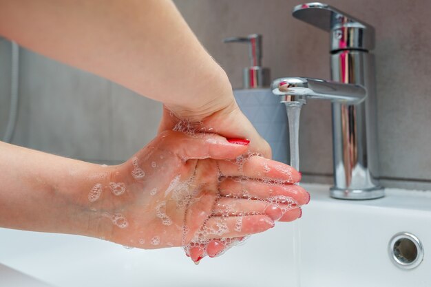 Lavabo blanco con grifo plateado en el baño. Lata gris con jabón líquido para manos. Abrir el agua del grifo, higiene personal de manos. Lavarse las manos con agua corriente