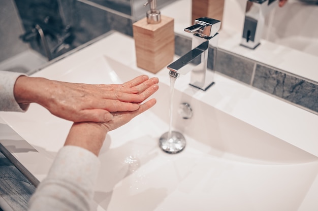 Lavado a mano espuma jabón líquido frotar las muñecas paso a mano mujer senior enjuague en agua en el lavabo del grifo del baño.