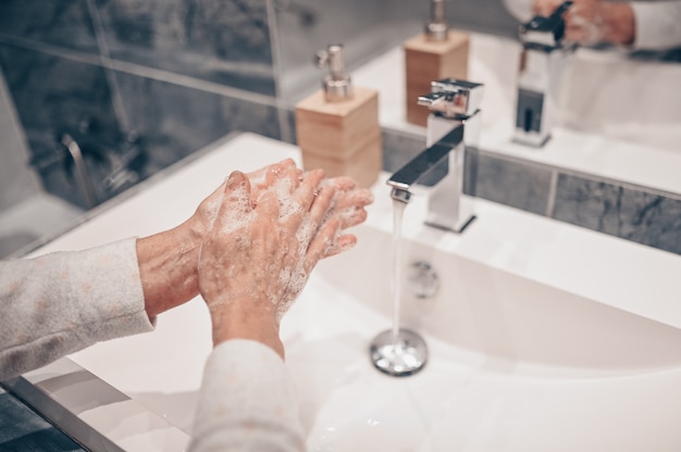 Lavado a mano espuma jabón líquido frotar las muñecas paso a mano mujer senior enjuague en agua en el lavabo del grifo del baño.