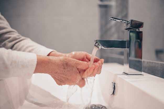 Lavado a mano espuma jabón líquido frotar las muñecas paso a mano mujer senior enjuague en agua en el lavabo del grifo del baño.