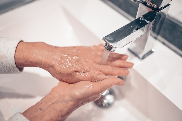 Lavado a mano espuma jabón líquido frotar las muñecas paso a mano mujer senior enjuague en agua en el lavabo del grifo del baño.