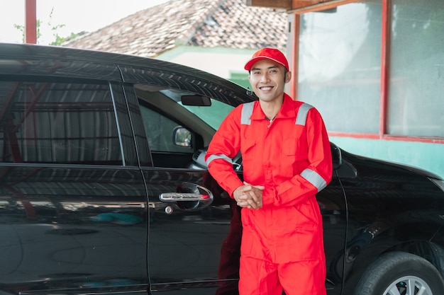 Lavadora de coches asiáticos vistiendo uniforme rojo sonriendo apoyado en el coche después de terminar el lavado de coches en el trabajo