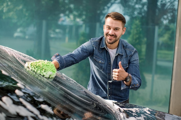 Lavagem de carros ao ar livre com esponja e espuma Lavagem de carros Homem barbudo sorrindo mostrando o polegar para cima limpando o para-brisa do carro usando esponja e espuma na estação de lavagem de carros ao ar livre