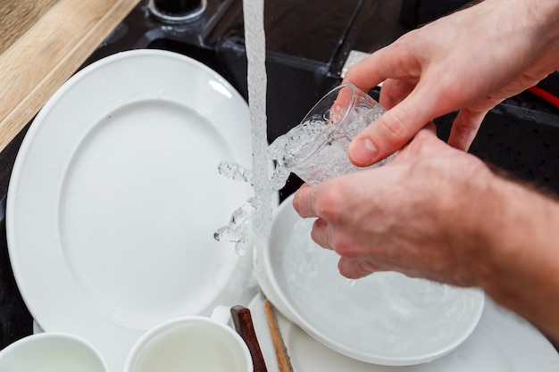Lavar los platos - las manos del hombre en los guantes de enjuagar el vaso con agua corriente en el fregadero.