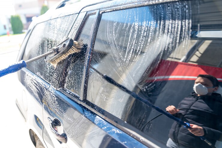 Foto lave a mano el automóvil con un cepillo con espuma. uso de la máscara durante el trabajo en el momento de la epidemia de coronavirus, reflejo en el cristal