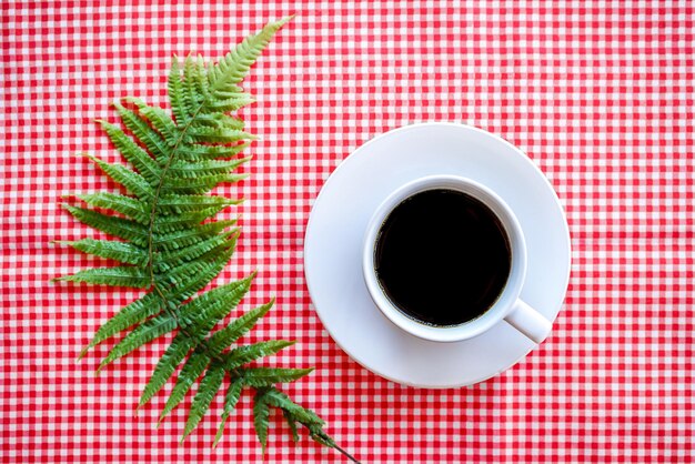 Lay Flat de café negro en taza de cerámica blanca con hojas de helecho verde sobre mantel rosa clásico