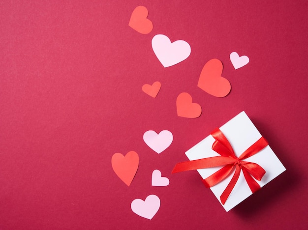 Lay Flat de celebración. Caja de regalo blanca con cinta roja sobre fondo rojo con corazones de papel. Concepto de San Valentín, día de la madre, día de la boda. Vista superior y espacio de copia.