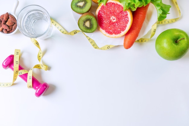 Lay Flat de frutas, verduras, mancuernas, cinta métrica y un vaso de agua en la mesa blanca