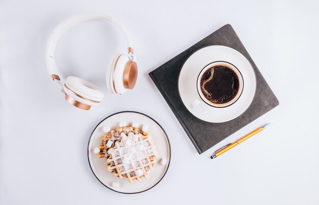 Lay Flat de gofres belgas con malvaviscos y café, auriculares y portátil sobre fondo blanco.