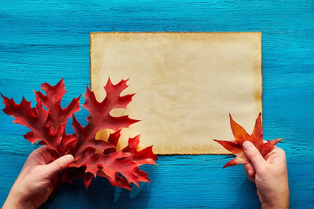 Lay Flat con página de papel envejecido y manos con hojas de otoño rojo vibrante en madera con textura turquesa. Colocar texto en papel.