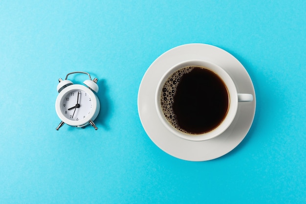 Foto layout criativo feito com uma xícara de café e despertador sobre fundo azul.