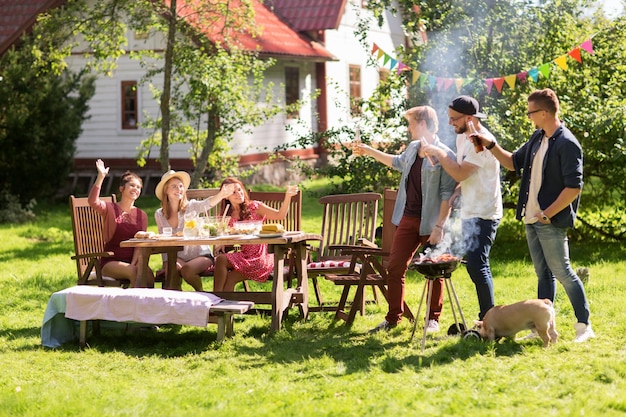 lazer, comida, pessoas, amizade e conceito de férias - amigos felizes cozinhando carne na churrasqueira e bebendo cerveja na festa ao ar livre de verão