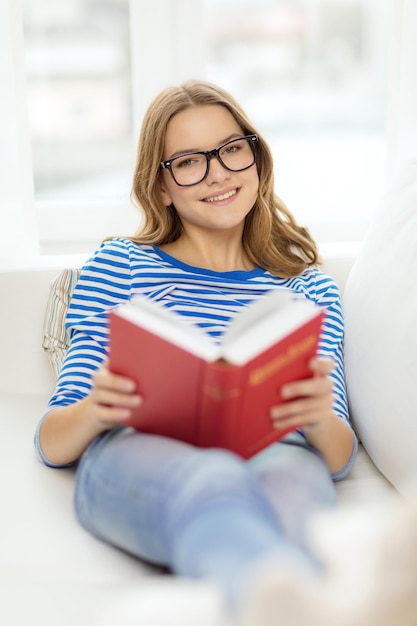 lazer, educação e conceito de casa - adolescente sorridente em óculos lendo livro e sentado no sofá em casa