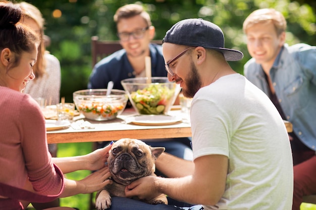 lazer, férias, alimentação, pessoas e conceito de comida - amigos felizes com cachorro bulldog francês jantando na festa no jardim de verão