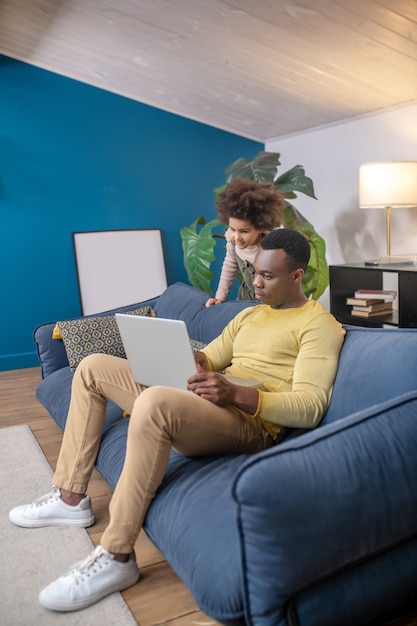 Foto lazer, laptop. pai adulto jovem de pele escura em roupas casuais com laptop sentado no sofá em casa e filhinha interessada atrás