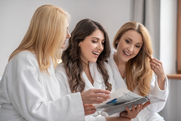 Lazer. Mulheres adultas bonitas e sorridentes com cabelos longos e jalecos brancos olhando uma revista e passando momentos de lazer juntas em uma sala iluminada