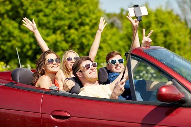 lazer, viagem, viagens e conceito de pessoas - amigos felizes dirigindo em cabriolet tirando fotos com bastão de selfie de smartphone no país