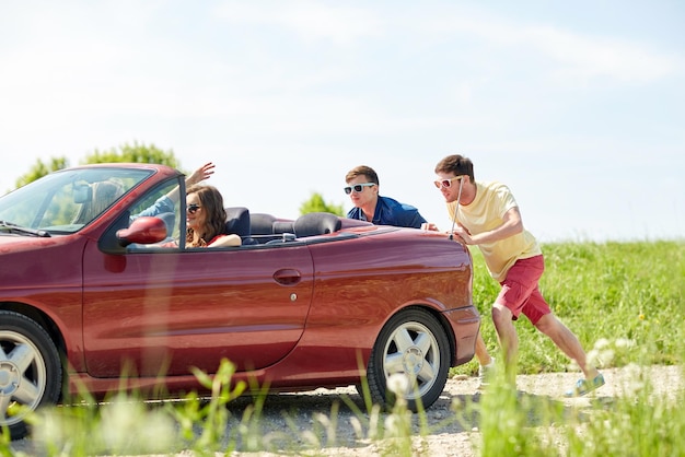 lazer, viagem, viagens e conceito de pessoas - amigos felizes empurrando carro cabriolet quebrado ao longo da estrada rural