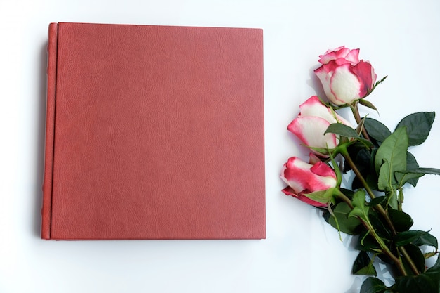 Álbum de boda cubierto de cuero rojo o libro de bodas y tres hermosas rosas se encuentra sobre fondo blanco.