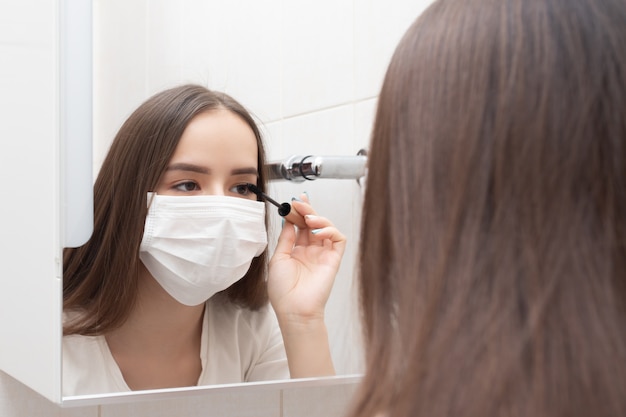 Leben in Quarantäne eines Coronovirus. Eine Frau in einer medizinischen Maske macht Make-up, Mascaras ihre Augen. Prävention und Schutz von Krankheiten