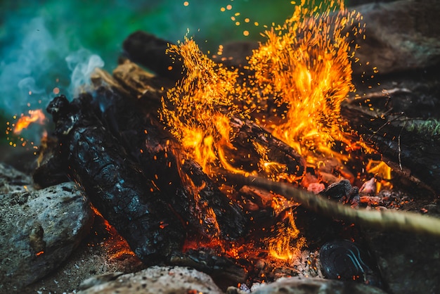 Lebendig schwelendes Brennholz brannte in Feuernahaufnahme. Atmosphärisch mit orangefarbener Flamme aus Lagerfeuer und blauem Rauch. Warmes Vollbild des Freudenfeuers. Glühende Glut in der Luft. Helle Funken im Bokeh