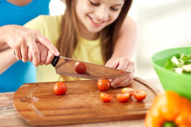 Lebensmittel-, Familien-, Koch- und Personenkonzept - Nahaufnahme eines glücklichen Mädchens und einer Mutter, die Abendessen zubereiten und Kirschtomaten auf Schneidebrett mit Messer in der Küche hacken