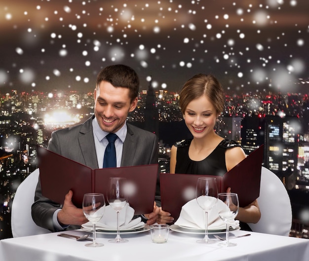 Lebensmittel-, Weihnachts-, Feiertags- und Personenkonzept - lächelndes Paar mit Menüs im Restaurant über verschneitem Nachtstadthintergrund