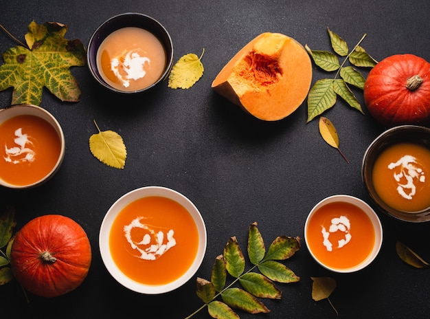 Lebensmittelhintergrund mit hausgemachter wärmender Kürbiscremesuppe in Tellern auf dunklem Tisch, dekoriert mit Kürbissen und gelben Blättern. Gesundes vegetarisches Essen des Herbstkomforts, Draufsicht, Kopienraum