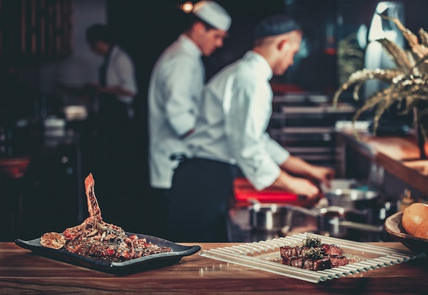 Lebensmittelkonzept. Fertig gegrilltes Schweinerippchengericht und Rindersteak mit Kräutern. Fertig zum Servieren. Fertig für den Verzehr. Zwei Köche, die im Hintergrund der modernen professionellen Restaurantküche arbeiten