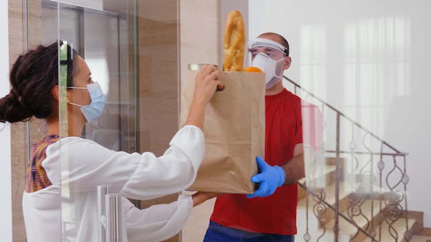 Lebensmittelkurier mit Schutzmaske, der einer Frau während Covid-19 Lebensmittel liefert.