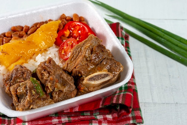 Lebensmittellieferung, Lunchpaket mit Rinderrippen gekocht mit Maniok, Reis, Bohnen und Tomaten auf weißem Holztisch.