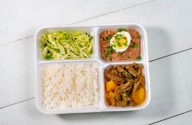 Lebensmittellieferung, Styropor-Lunchbox mit Rindfleisch, gekocht mit Maniok, Zucchini, Reis, Bohnen und gekochtem Ei auf weißer Holzstruktur. Ansicht von oben.