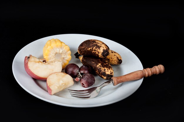 Lebensmittelverschwendungs-, Apfel-, Bananen-, Mais- und Traubenfrucht mit Gabel auf Teller