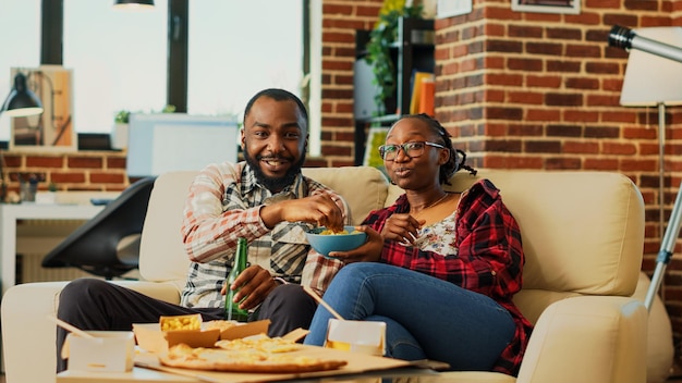 Lebenspartner trinken Alkohol und essen Snacks, während sie sich im Fernsehen Comedy-Filme ansehen. Junger Mann und Frau bestellen Essen zum Mitnehmen und schauen sich Filme im Fernsehen an und haben Spaß. Stativaufnahme.