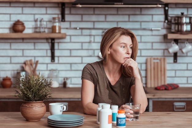 Lebensprobleme. Depressive Frau, die in der Küche sitzt, Tabletten nimmt und über Lebensprobleme nachdenkt