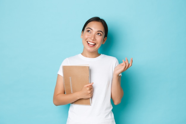 Lebensstil, Bildung und Menschenkonzept. Glückliche schöne asiatische Studentin im weißen T-Shirt, Mädchen, das verträumte und suchen verträumte obere linke Ecke, während Notizbücher, hellblaue Wand hält