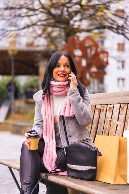 Lebensstil, ein kaukasisches brünettes Mädchen, das in der Stadt mit Papiertüten und einem Kaffee zum Mitnehmen einkauft und auf einem Anruf sitzt