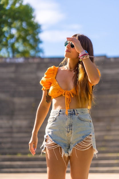 Lebensstil einer jungen kaukasischen Brünette, die im Sommer in einem Park in der Stadt spaziert. Mädchen mit Sonnenbrille und orangefarbenem T-Shirt