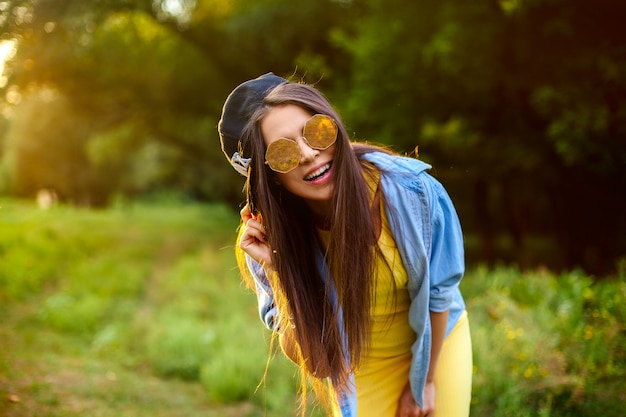 Lebensstil. glückliches Mädchen in der Sonnenbrille und in einer Kappe in der hellen Kleidung in einem Park bei Sonnenuntergang