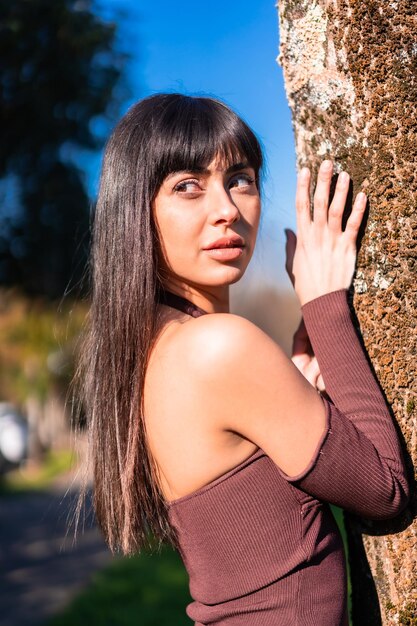 Lebensstil im Frühling. Posieren eines brünetten Modelmädchens, das sich mit einem braunen Kleid an einen Baum in einem Park lehnt und nach links schaut
