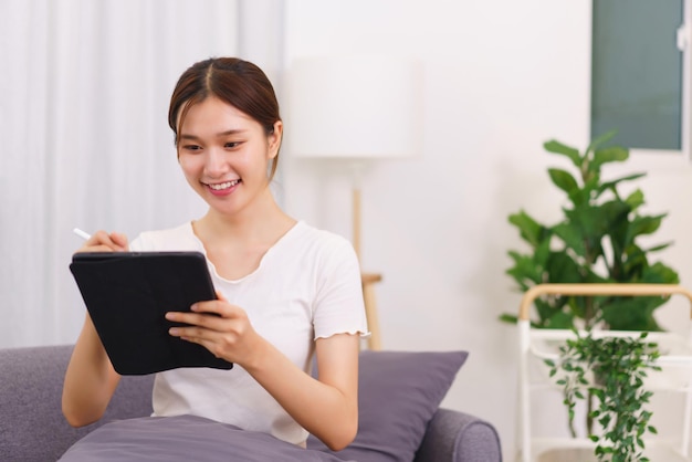 Lebensstil im Wohnzimmerkonzept Junge asiatische Frau, die Daten auf dem Tablet schreibt, während sie auf der Couch sitzt