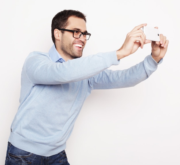 Lebensstil, Technologie und Menschenkonzept: Ein junger Mann im Hemd, der ein Mobiltelefon hält und ein Foto von sich selbst macht, während er vor weißem Hintergrund steht.