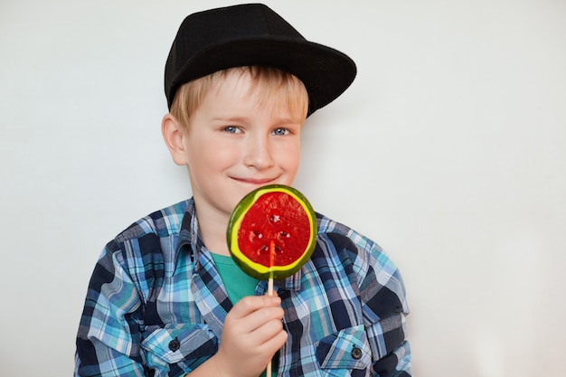 Lebensstil und Menschenkonzept. Entzückendes kleines männliches Kind mit blauen Augen und hellem Haar in der schwarzen Kappe und im karierten Hemd, das mit Süßigkeit aufwirft