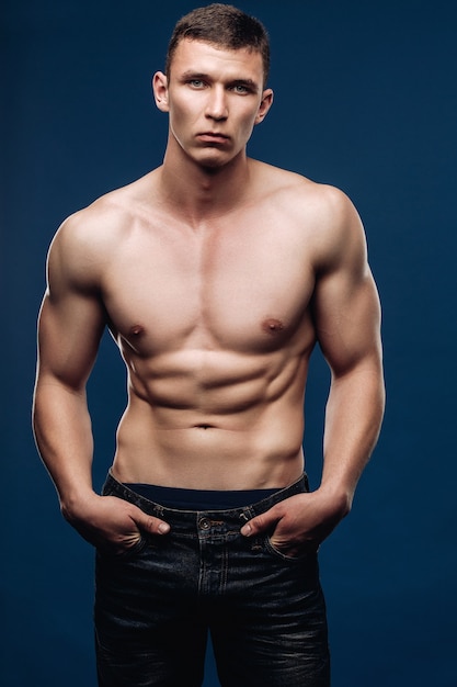 Lebensstilporträt eines männlichen Modells mit einem muskulösen Torso, der die Kamera betrachtet, Hände in der Tasche der Jeans, Hose oben. Der blaue Hintergrund des Studios.