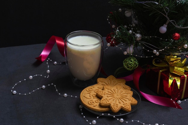 Lebkuchen und Milch für den Weihnachtsmann. Weihnachtskomposition mit Lebkuchen und Milch auf schwarzem Hintergrund mit Weihnachtsbaum und Geschenk, Draufsicht