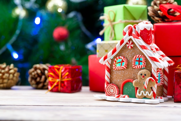 Lebkuchenhaus auf einem Holztisch zusammen mit Weihnachtsgeschenken auf dem eines Weihnachtsbaumes.