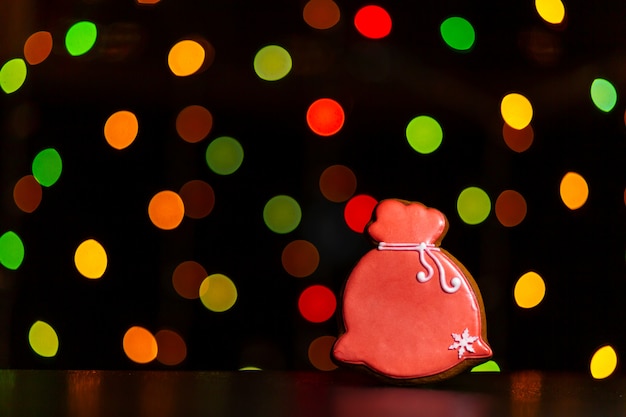 Lebkuchenplätzchen der roten Tasche Sankt mit Geschenken über defocused farbigen Lichtern der Girlande.