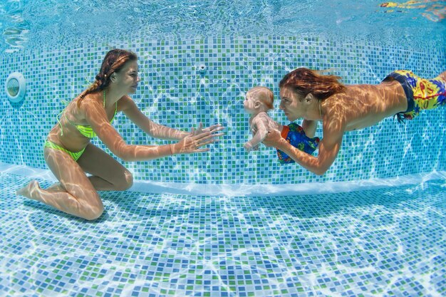 Lección de natación infantil: bebé con madre, padre aprendiendo a nadar, buceando bajo el agua en la piscina.